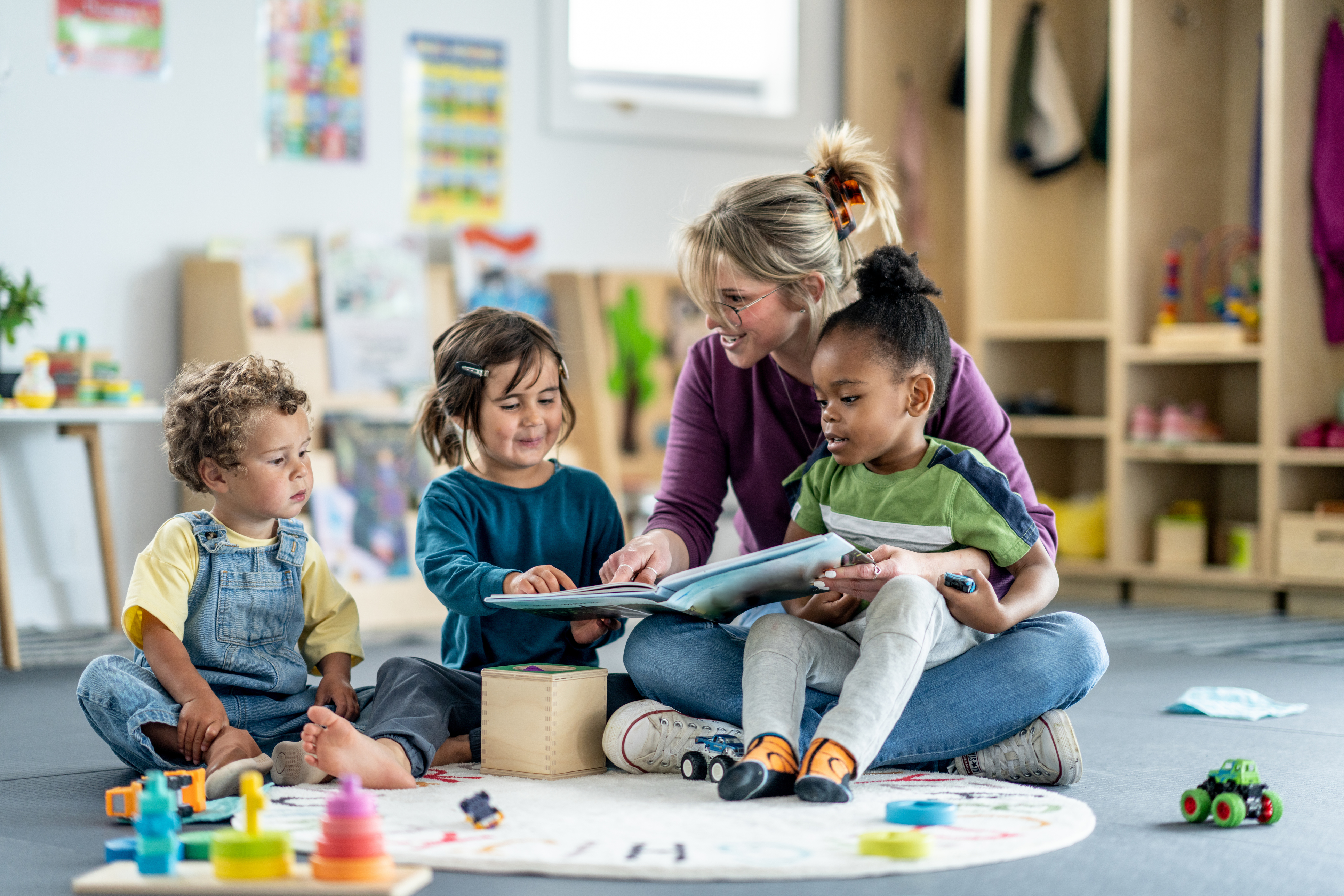 A group of children in a daycare or classroom setting engages with a teacher reading a book to them